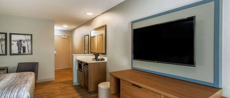 A modern hotel room with a bed, a TV mounted on the wall, a wooden dresser, a small trash bin, and a bathroom with a sink and mirror.
