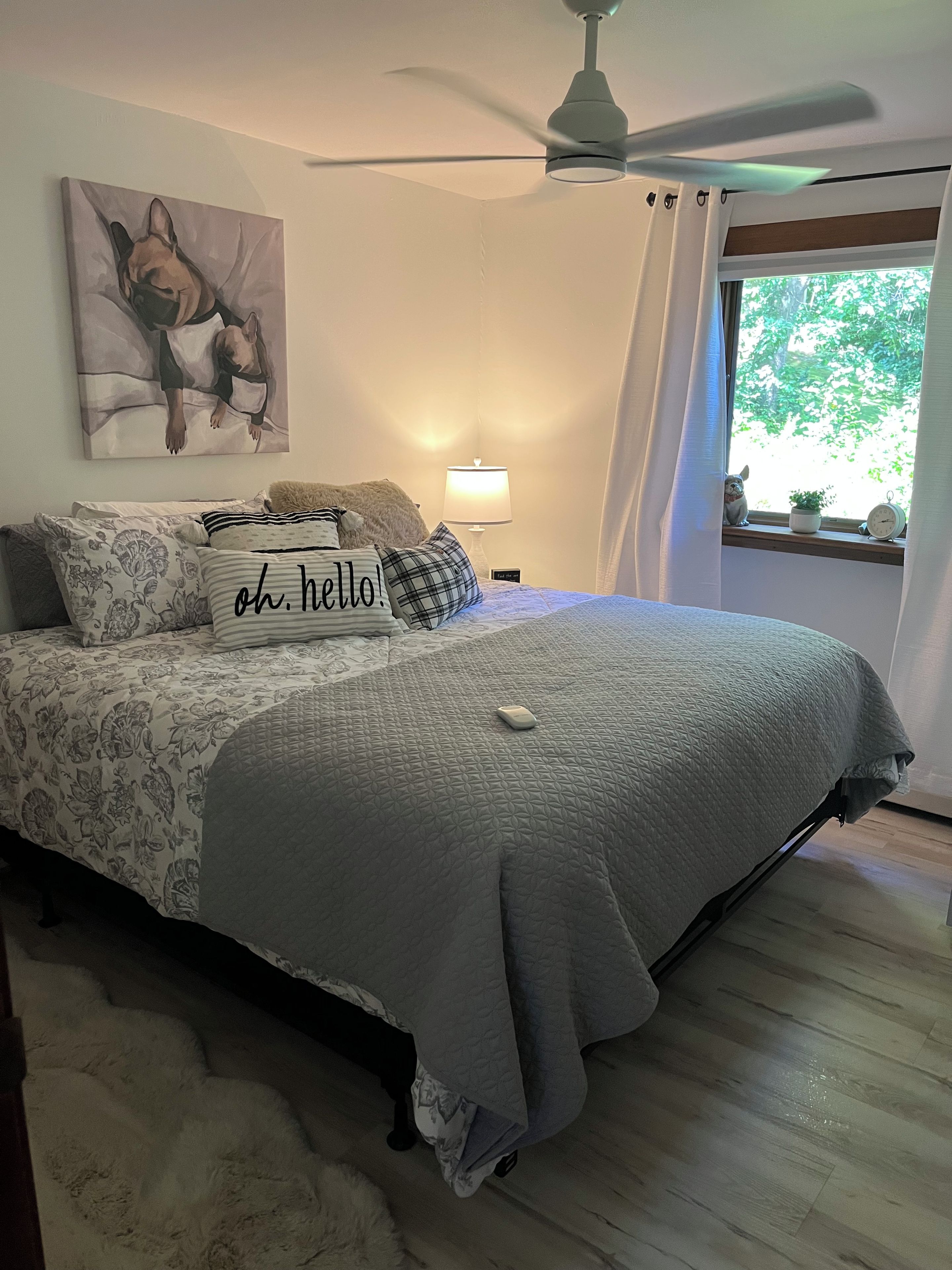 A bedroom with a bed, a ceiling fan, a window with curtains, a bedside lamp, and a painting on the wall.