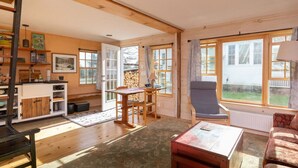 A spacious living room with a wooden floor, a dining area with a table and chairs, a sofa, and a large window offering a view of the outside. - Absolutely Gorgeous Vermont Garden Cottage (Montpelier)