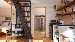 A cozy cabin interior with a wooden staircase leading to a loft bedroom, a kitchen with modern appliances, and a bookshelf filled with books. - Absolutely Gorgeous Vermont Garden Cottage (Montpelier)