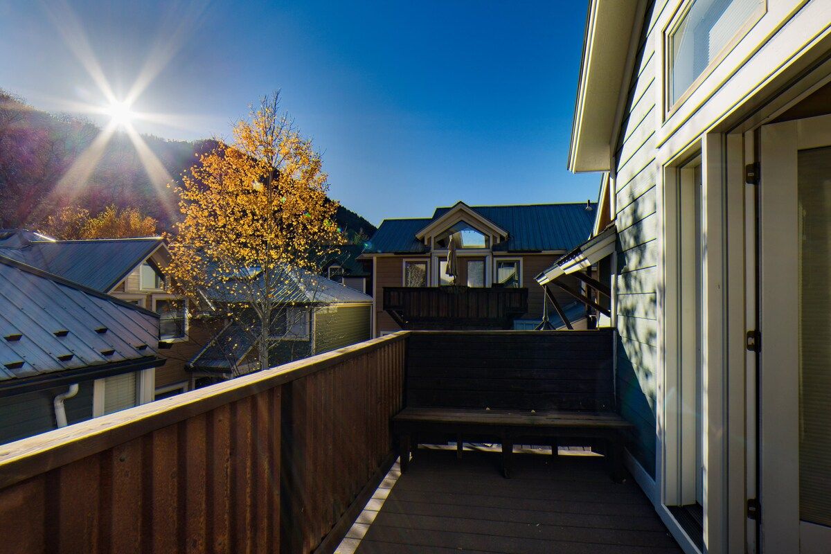 A wooden deck with a bench, a house with a sloped roof, and a tree with autumn leaves.