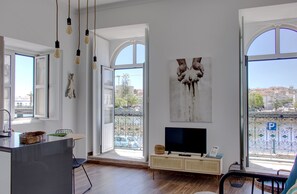 Een moderne woonkamer met een grote televisie, open ramen en een balkon met uitzicht op de stad.
