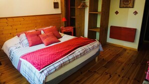 Een slaapkamer met een houten hoofdeinde, een bed met rood-wit beddengoed, een rode deken en een houten garderobe met spiegel.