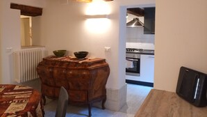 Een eethoek met een houten tafel en stoelen, een houten buffetkast met twee kommen, een radiator, en een keuken zichtbaar op de achtergrond.
