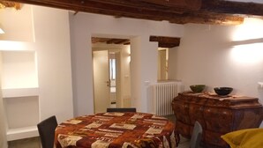 Een eethoek met een ronde tafel, bedekt met een quilt, omringd door stoelen. Achtergrond toont een houten kastje met een kom erop.