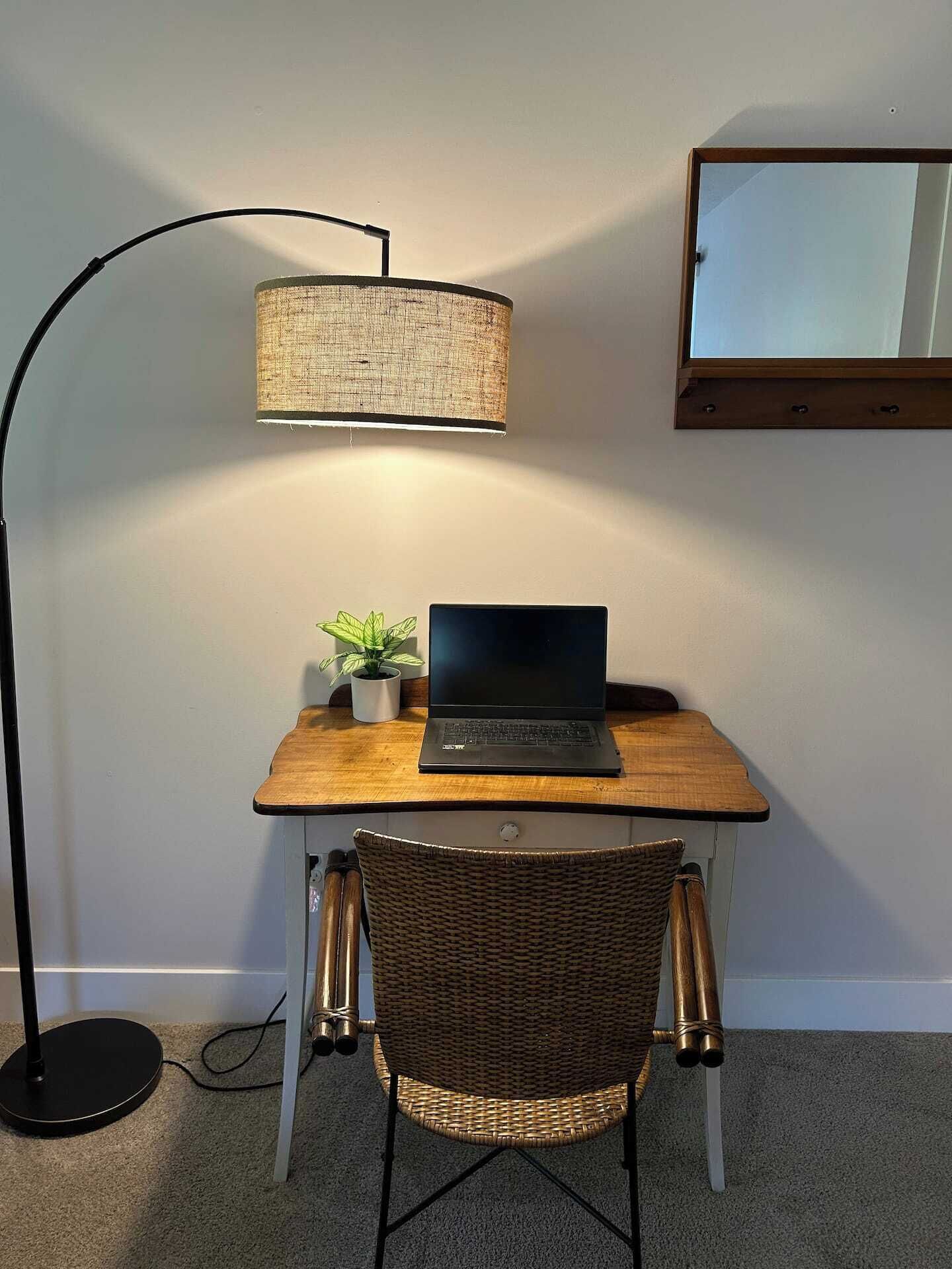 Um espaço de trabalho com um laptop sobre uma escrivaninha de madeira, uma luminária de chão, uma cadeira de vime e uma pequena planta em vaso.