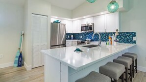 A modern kitchen with white cabinets, a stainless steel refrigerator, a microwave, and a blue-tiled backsplash. There are two bar stools and a large island with a sink.