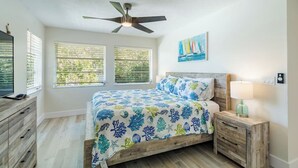 A bedroom with a bed, a ceiling fan, a nightstand with a lamp, and a painting on the wall.
