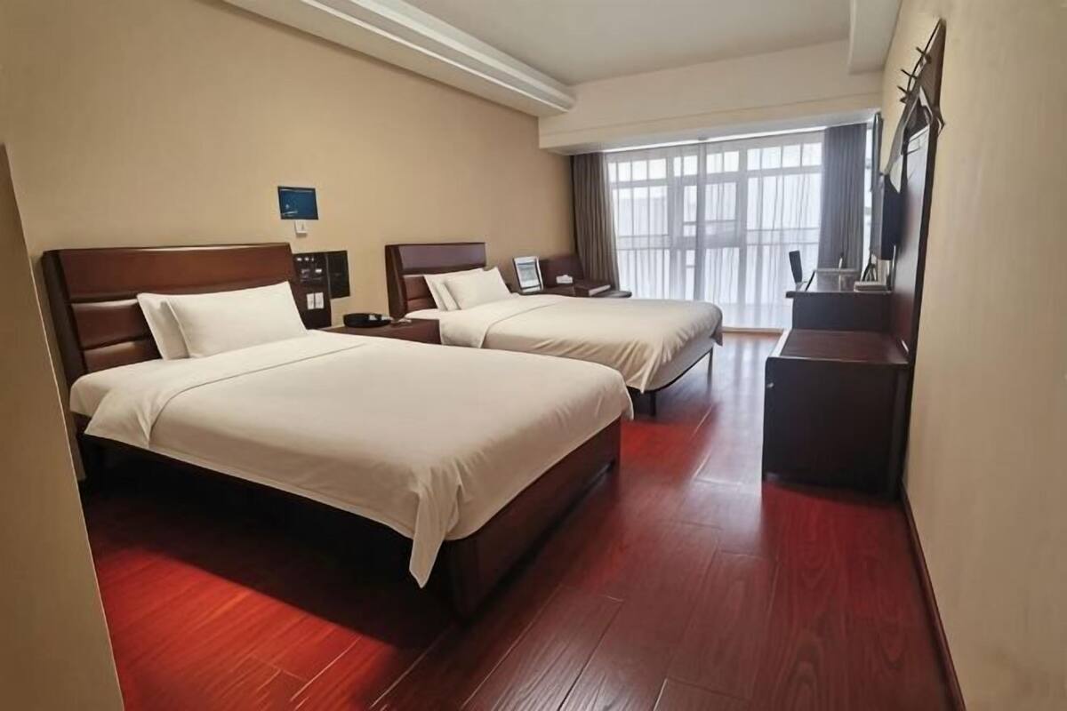 A hotel room with two beds, a desk, a nightstand, and a large window.