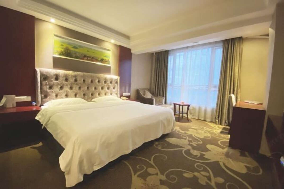 A hotel room with a large bed, a chair, a small table, and a painting on the wall.