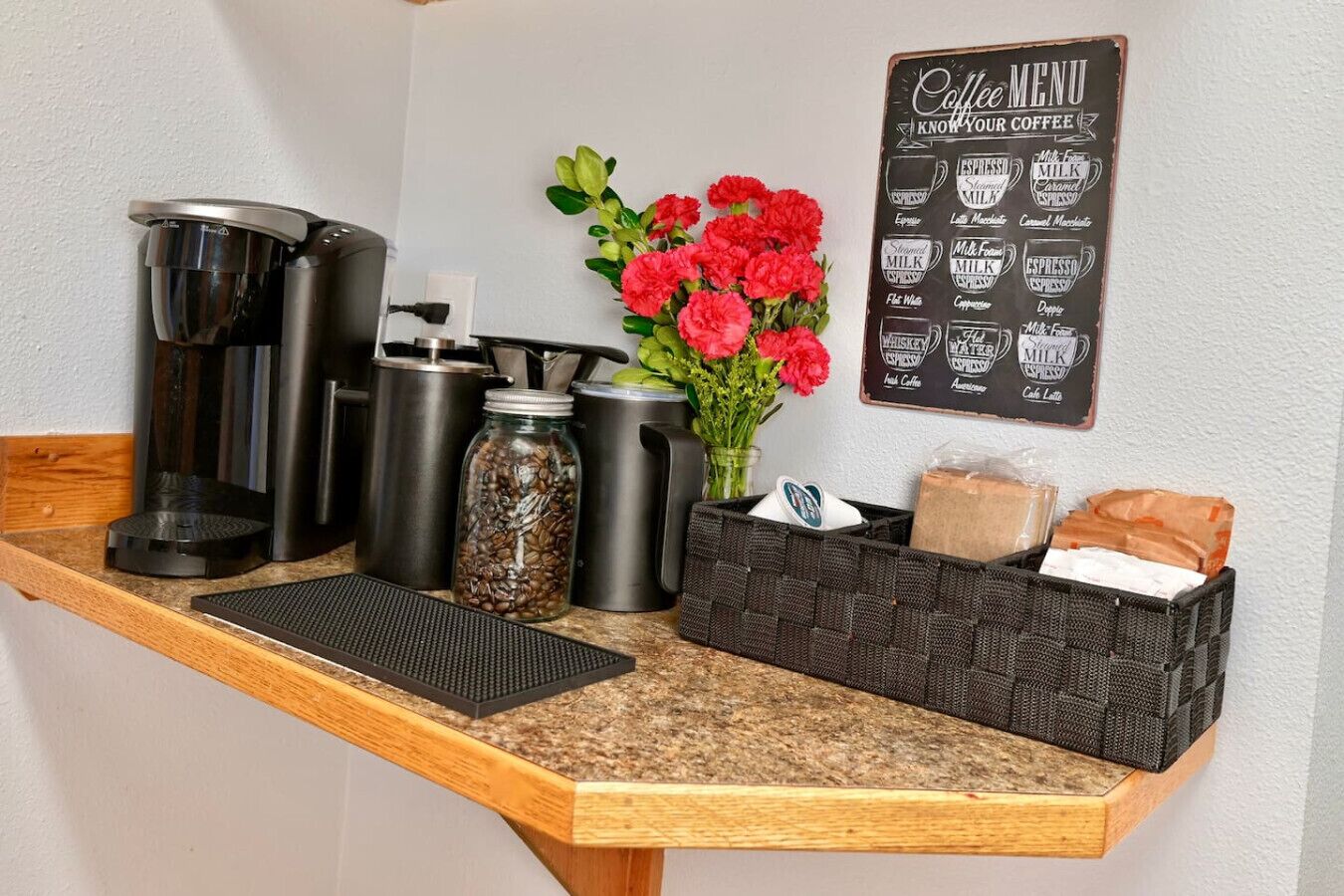 A coffee station with a coffee menu, coffee makers, a jar of coffee beans, a vase with flowers, and a basket of bread.