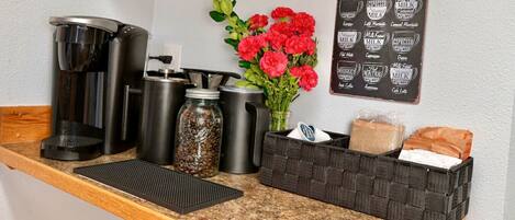 A coffee station with a coffee menu, coffee makers, a jar of coffee beans, a vase with flowers, and a basket of bread.