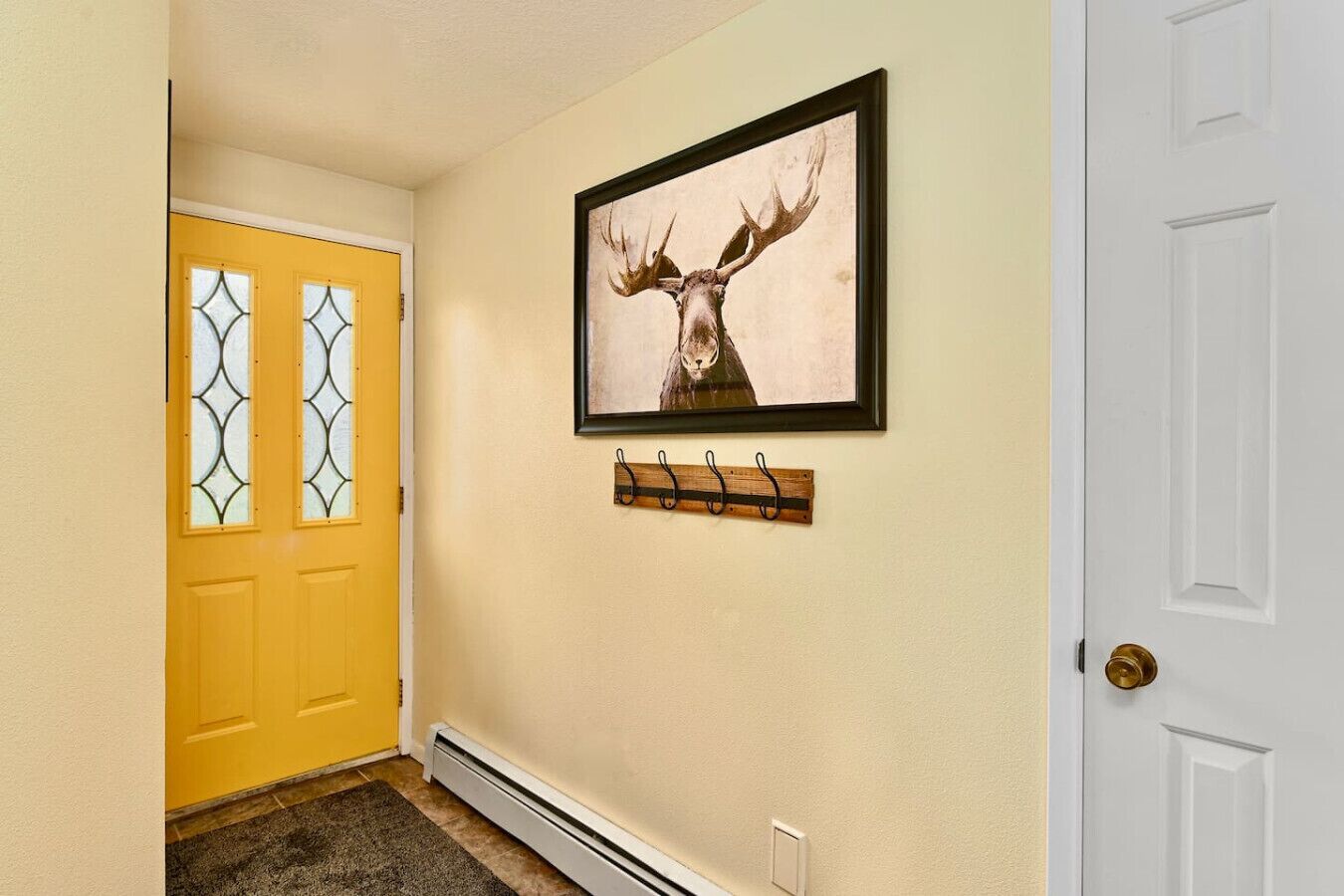 A hallway with a yellow door, a white door, and a framed deer head picture on the wall.
