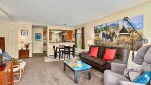 A living room with a brown leather sofa, a coffee table, and a dining area with a table and chairs. - Sneaky Moose Townhome, 2BR / 2BA (Kenai)