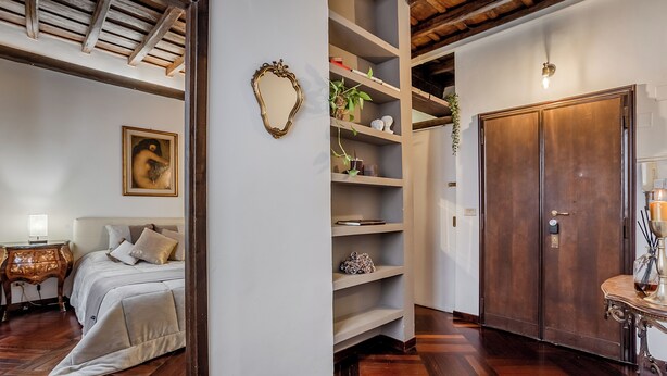 Un dormitori amb un llit, una taula de nit i un mirall. Hi ha una porta de fusta i una prestatgeria amb objectes decoratius.