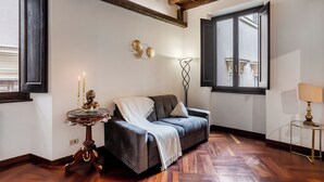 Una habitació acollidora amb un sofà gris fosc, una taula rodona petita amb un objecte decoratiu, una làmpada de peu i una finestra amb vista a edificis.