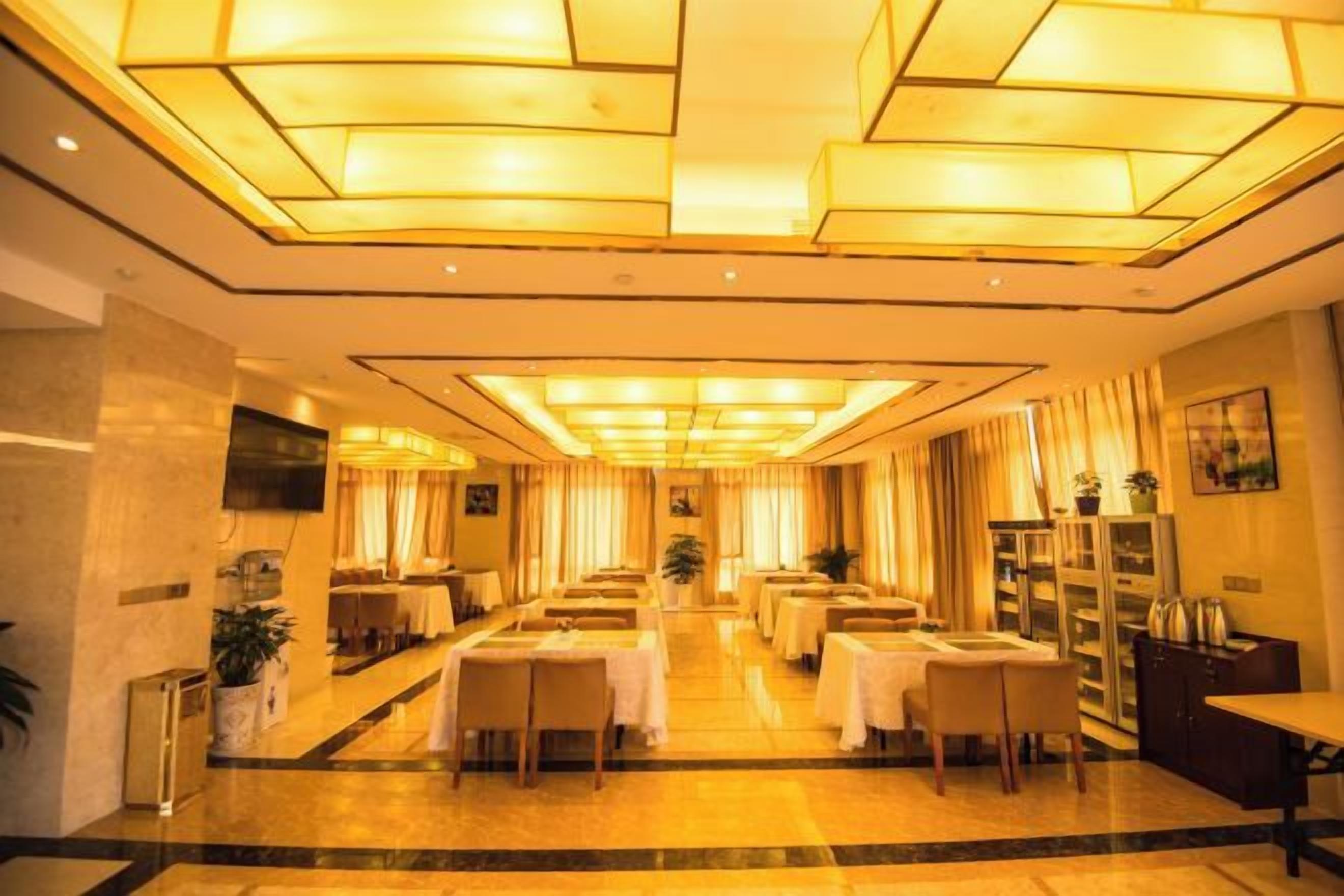 Comedor de hotel con varias mesas y sillas, un candelabro central y un televisor instalado en la pared.