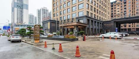 Entrada de un hotel con un edificio grande al fondo, pavimento mojado y conos de tráfico.