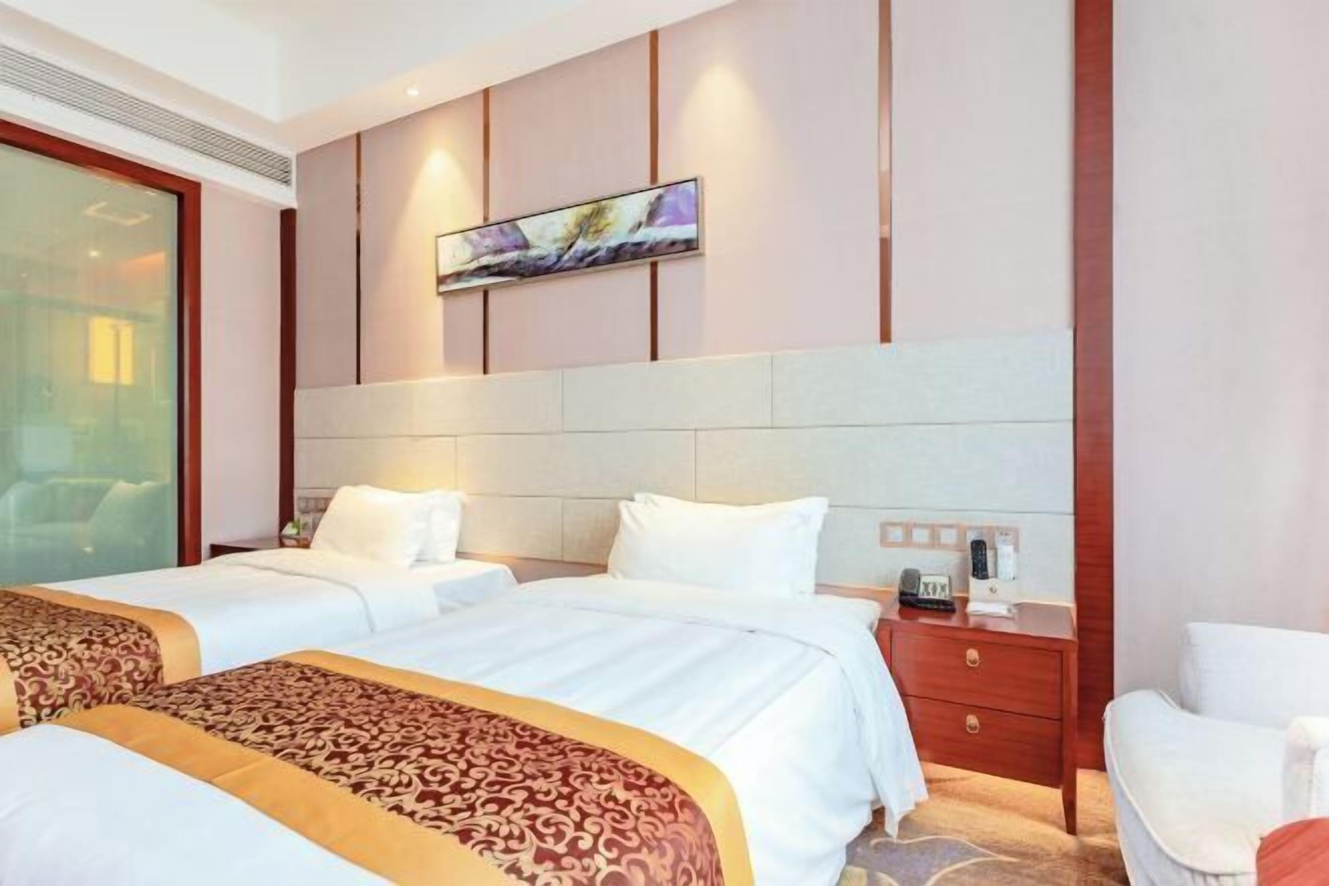 A hotel room with two beds, a large window, a painting on the wall, and a bedside table with a lamp.