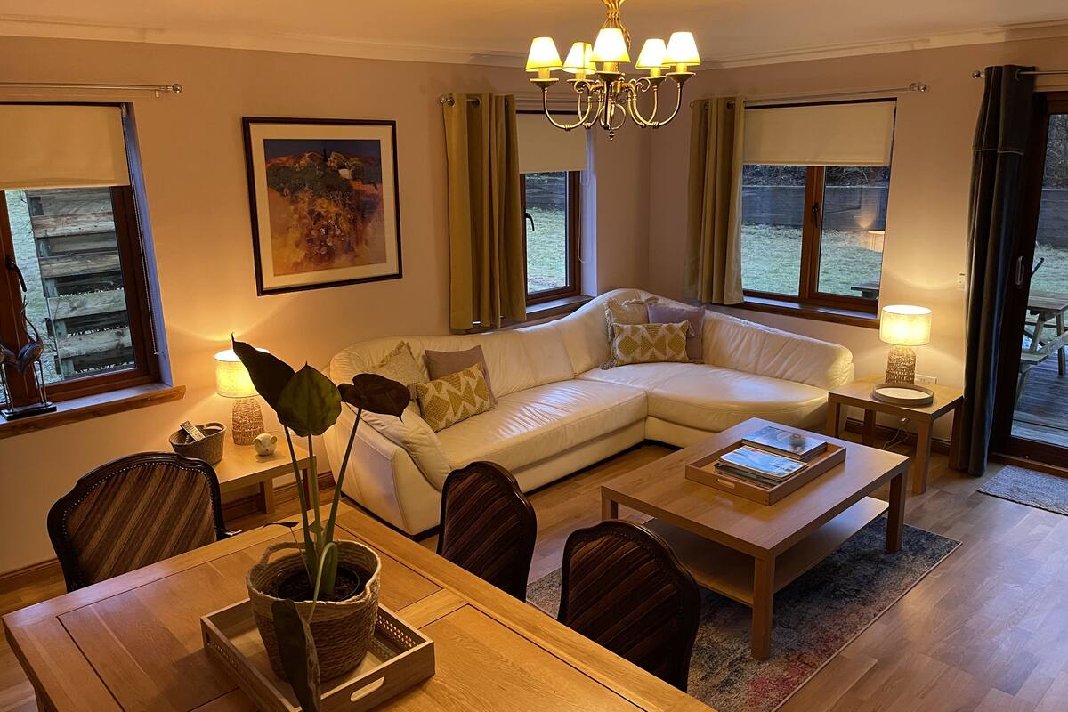 A living room with a white sectional sofa, a wooden coffee table, a dining table with chairs, a chandelier, and large windows with curtains.