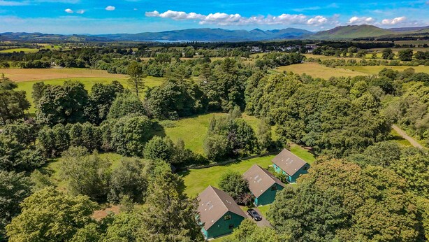 An aerial view of a rural area with two green-roofed buildings surrounded by dense forests and open fields. - Foxglove Cottages (Drymen)