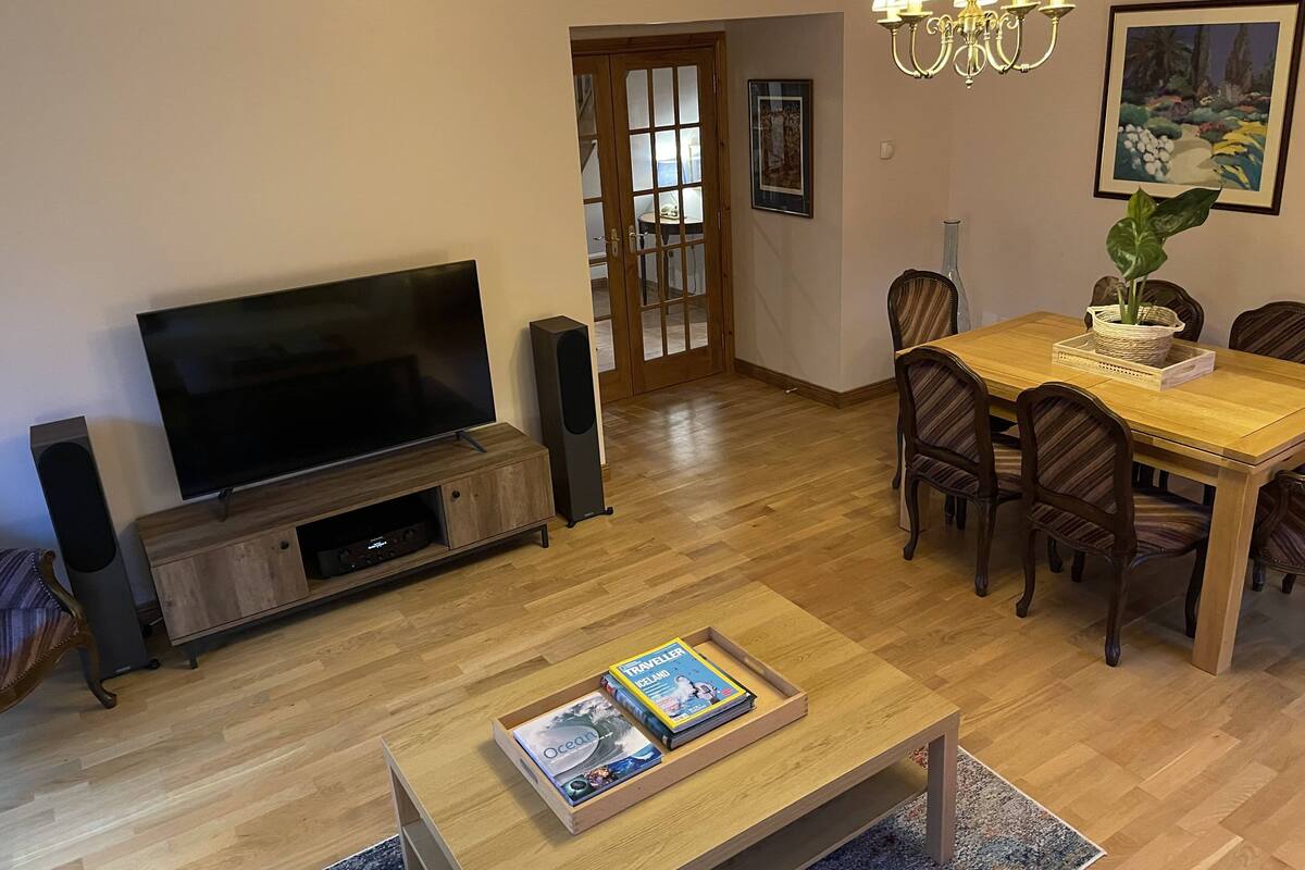 A living room with a TV, speakers, a coffee table with books, a dining table with chairs, a chandelier, and a door leading to another room.