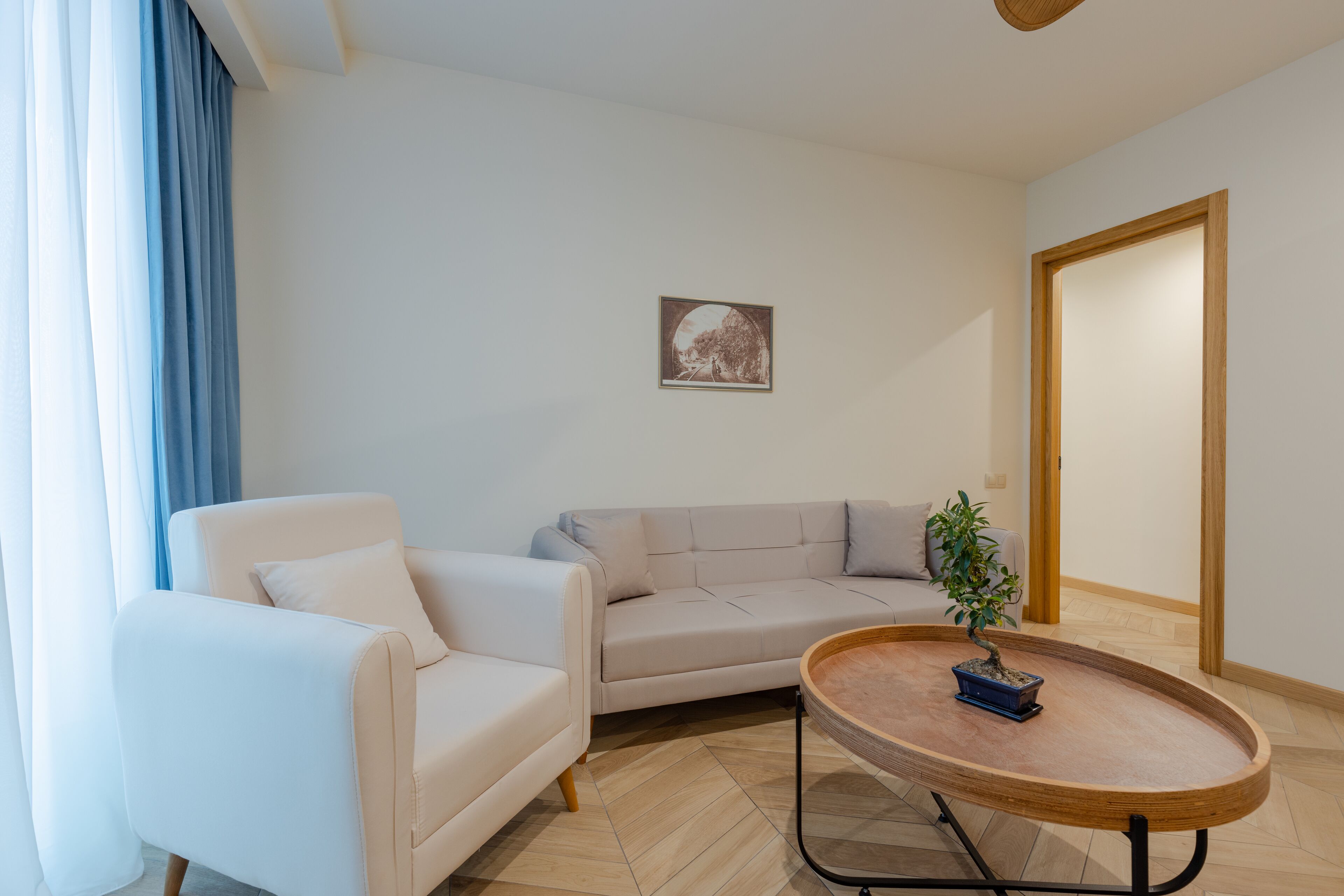 Una sala de estar moderna con un sofá color beige, un sillón blanco, una mesa redonda de centro con un bonsái y un cuadro en la pared.