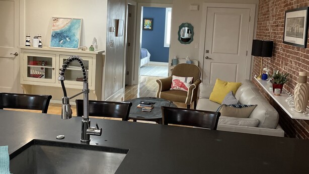 A modern kitchen with a black countertop, white cabinets, and a dining area with a table and chairs. There is a brick wall and a blue door in the background. - Entire condo center of downtown Newburyport (Newburyport)