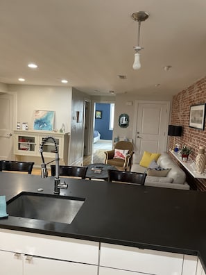 Une cuisine moderne avec un plan de travail noir, des armoires blanches et un coin repas comprenant une table et des chaises. On aperçoit un mur en briques et une porte bleue à l’arrière-plan.