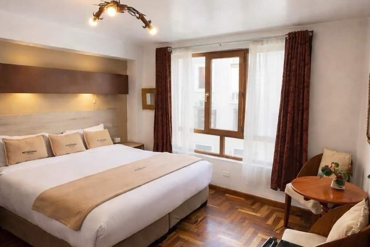 A hotel room with a large bed, a desk, a chair, a small table, a window with curtains, and a ceiling light fixture.