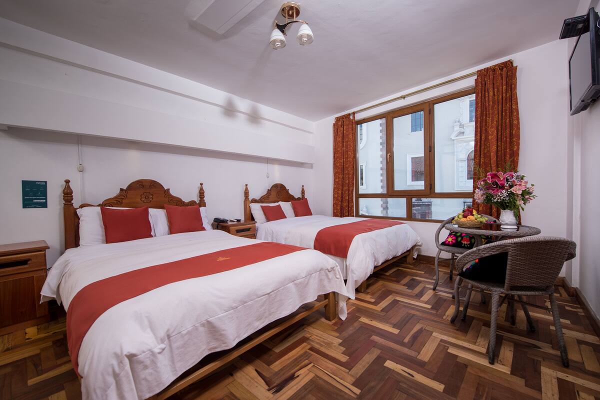 A hotel room with two beds, a wooden floor, a window with curtains, a small table with a vase, and a television on the wall.