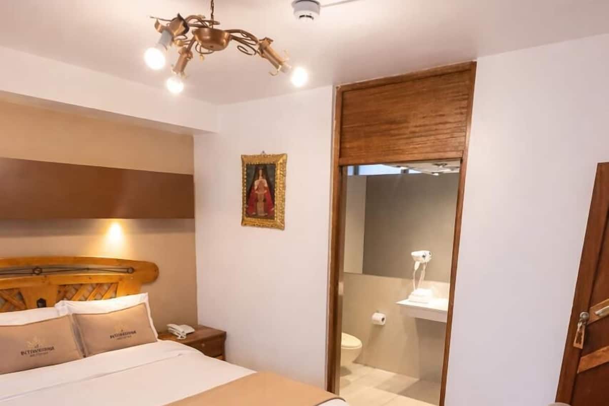 A hotel room with a wooden headboard, a bed with a white bedspread, a wooden floor, a small bedside table with a lamp, and a door leading to a bathroom.
