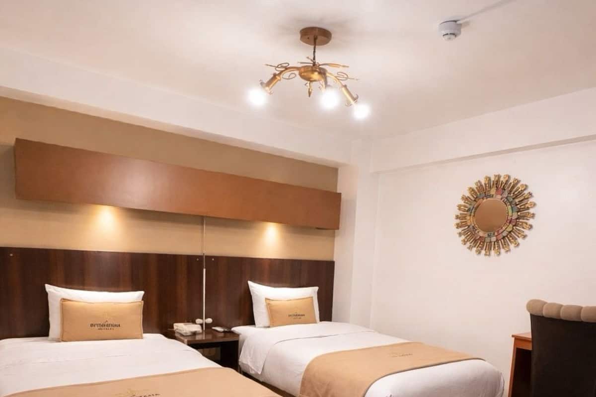 A hotel room with two beds, a wooden herringbone floor, a ceiling light fixture, and a decorative wall piece.