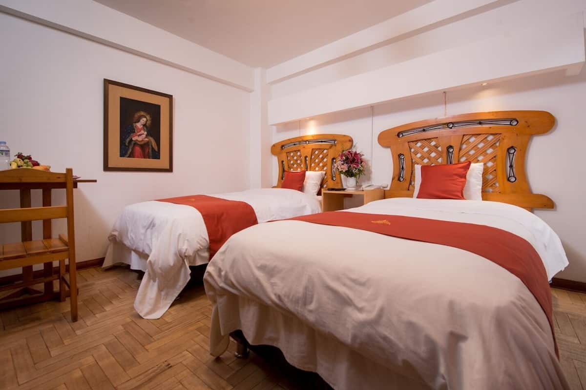 A hotel room with two beds, a wooden headboard, a wooden chair, a framed picture on the wall, and a small table with a vase of flowers.