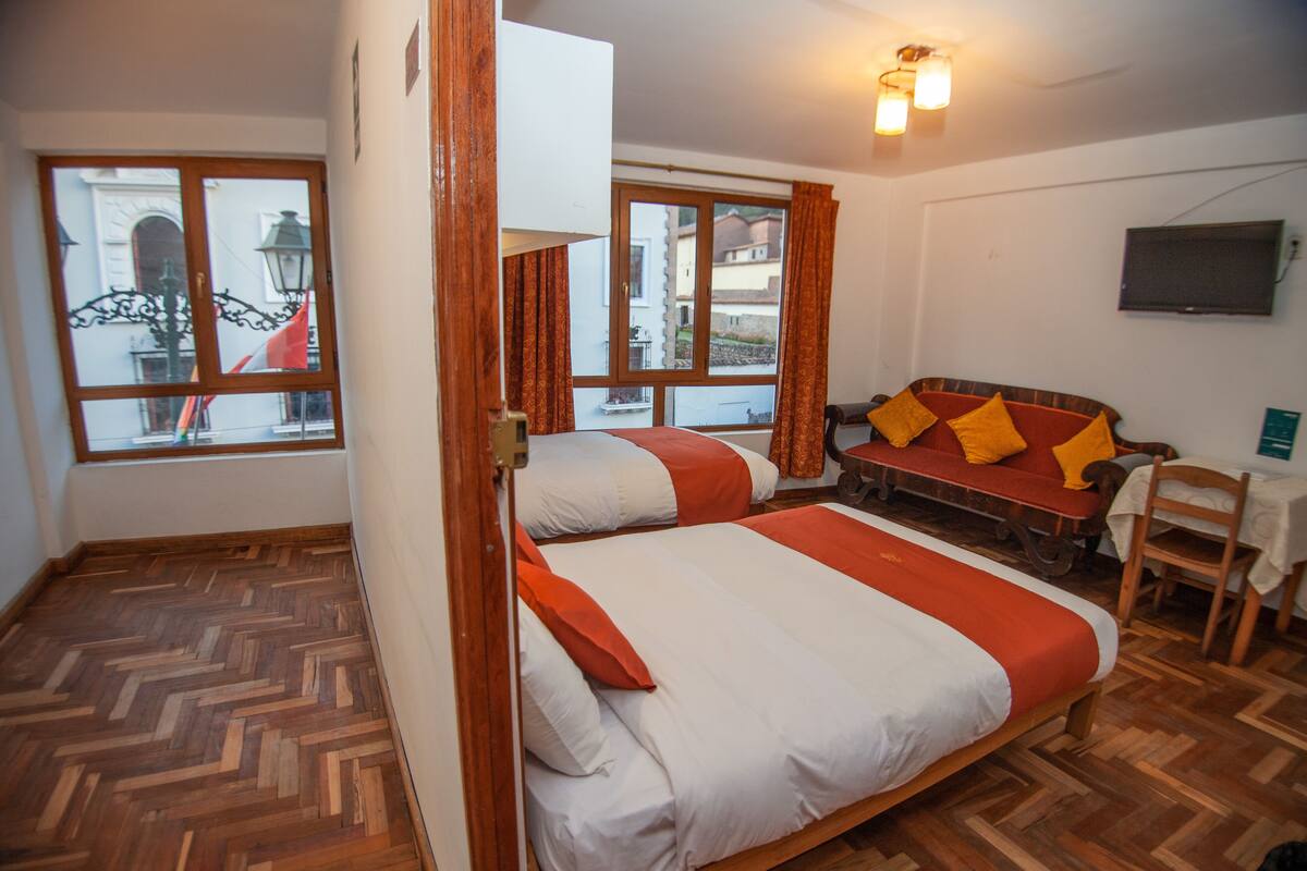 A room with two beds, a wooden floor, a sofa with cushions, a chair, and a TV on the wall.