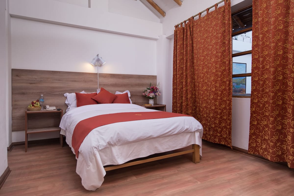 A bedroom with a wooden headboard, a bed with white and red bedding, a bedside table with a lamp, a nightstand with a phone and a basket of fruit, and a window with curtains.