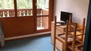 A room with a wooden desk, a chair, a television, and a view of a forest through large windows. - Studio terrace with beautiful view and direct access to all hiking trails; sleeps 4 (Modane)