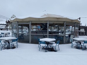 Un área de estar al aire libre cubierta de nieve, con mesas y sillas, un cerramiento de vidrio y un letrero que indica que el establecimiento está cerrado.
