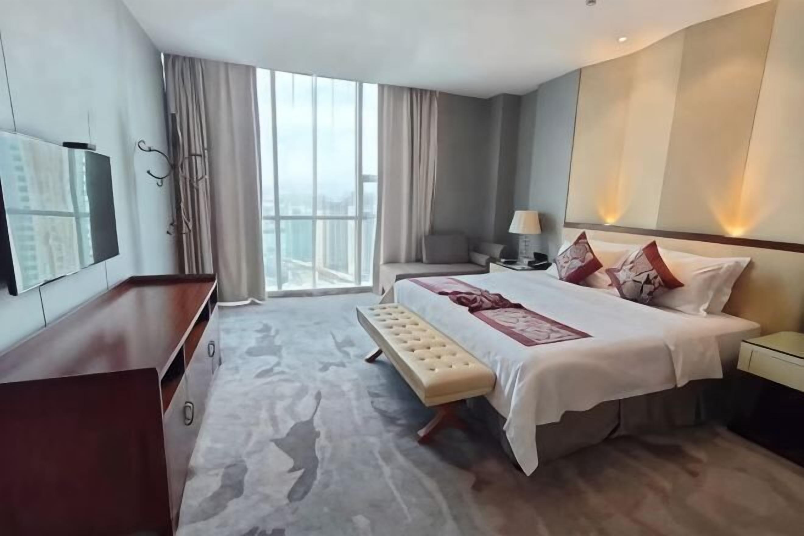 Habitación de hotel con una cama grande, un banco, un escritorio, un televisor y vistas de la ciudad.