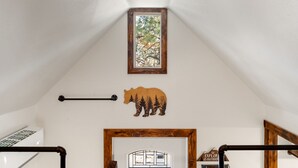Een kamer met schuine plafond, een raam met uitzicht op bomen, een houten beren decoratie, een lijst met afbeelding en een houten deur met een bordje.