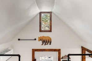 Une pièce avec un plafond en pente, une fenêtre donnant sur des arbres, une décoration en forme d’ours en bois, un tableau encadré et une porte en bois avec une inscription.