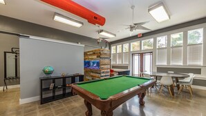 A pool table in a room with a ceiling fan, a globe on a console table, and a large window with blinds.