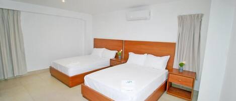 Habitación de hotel con dos camas individuales, cabecera de madera, mesita de noche con lámpara y un cajón pequeño.