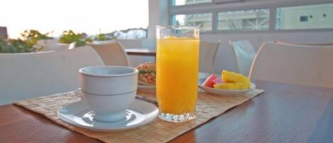 A breakfast setting with a glass of orange juice, a cup, and a plate of fruit on a table with a view of buildings through a window.