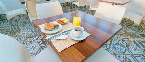 Et spiseområde med et træbord, hvide stole, en tallerken med mad, et glas appelsinjuice og en kop kaffe.