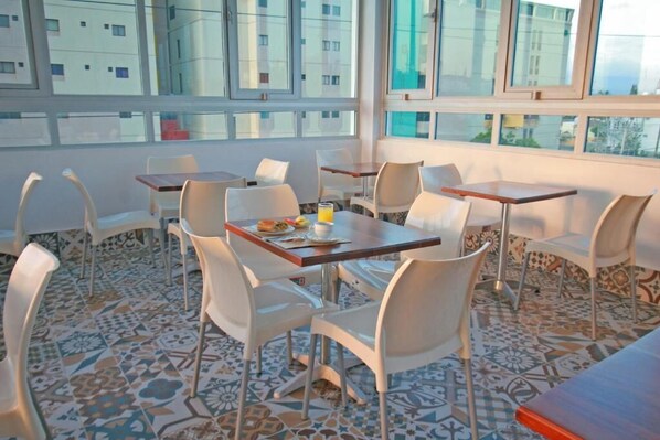 Un espace repas avec des tables et des chaises, de grandes fenêtres et un sol carrelé.