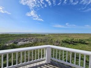 Una terraza de madera con barandillas blancas que da vista a un paisaje costero con zonas verdes y vistas al océano.