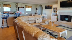 Un salon comprenant un canapé d’angle en cuir, une table basse en bois et un téléviseur fixé au-dessus d’une cheminée. On y trouve également un coin cuisine avec un réfrigérateur, une table à manger et des tabourets de bar.