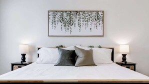 Una cama bien hecha con un cabecero de madera, dos mesitas de noche con lámparas y una obra de arte enmarcada en la pared.