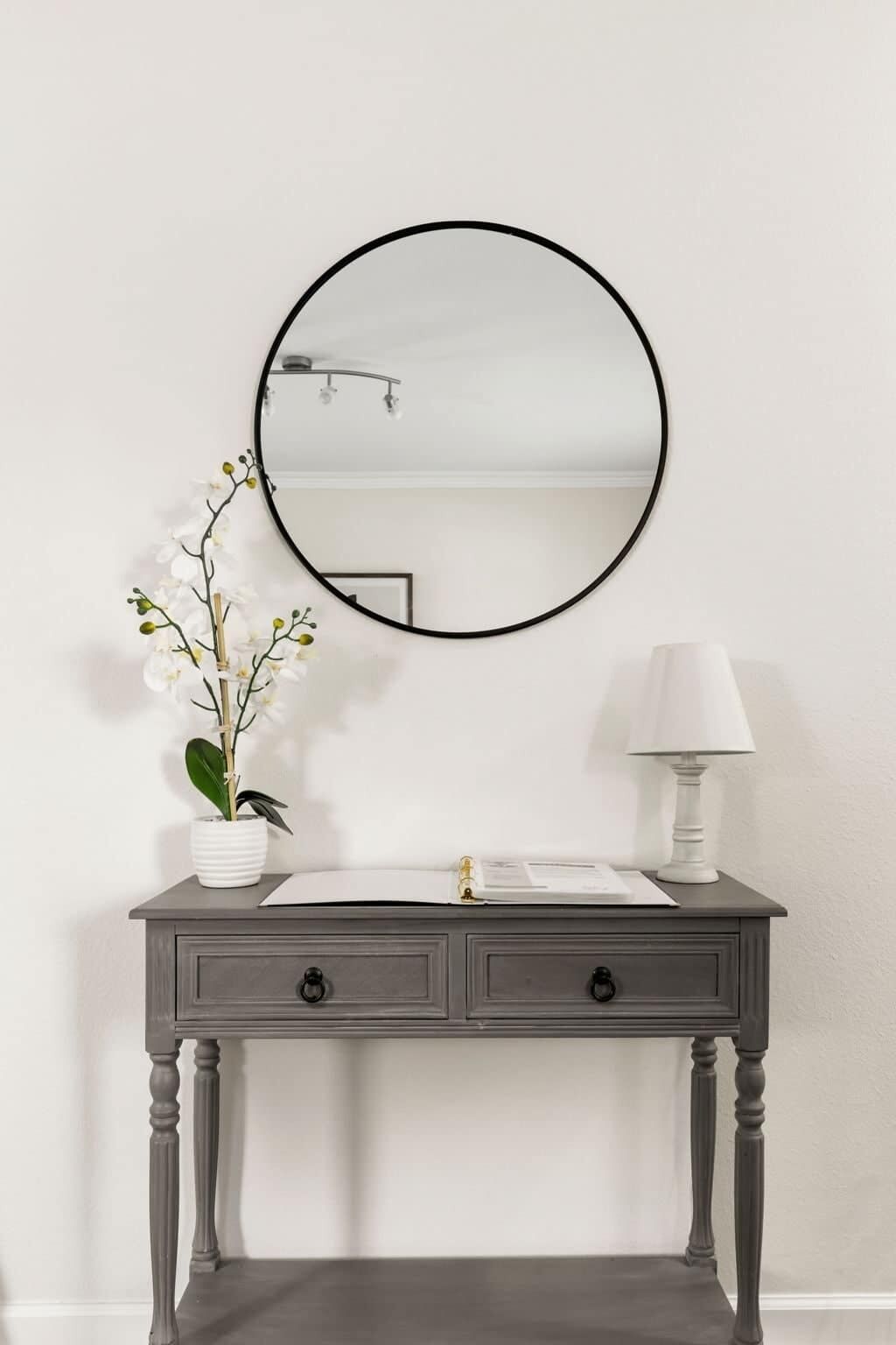 A vanity with a round mirror, a white vase with flowers, and a lamp.